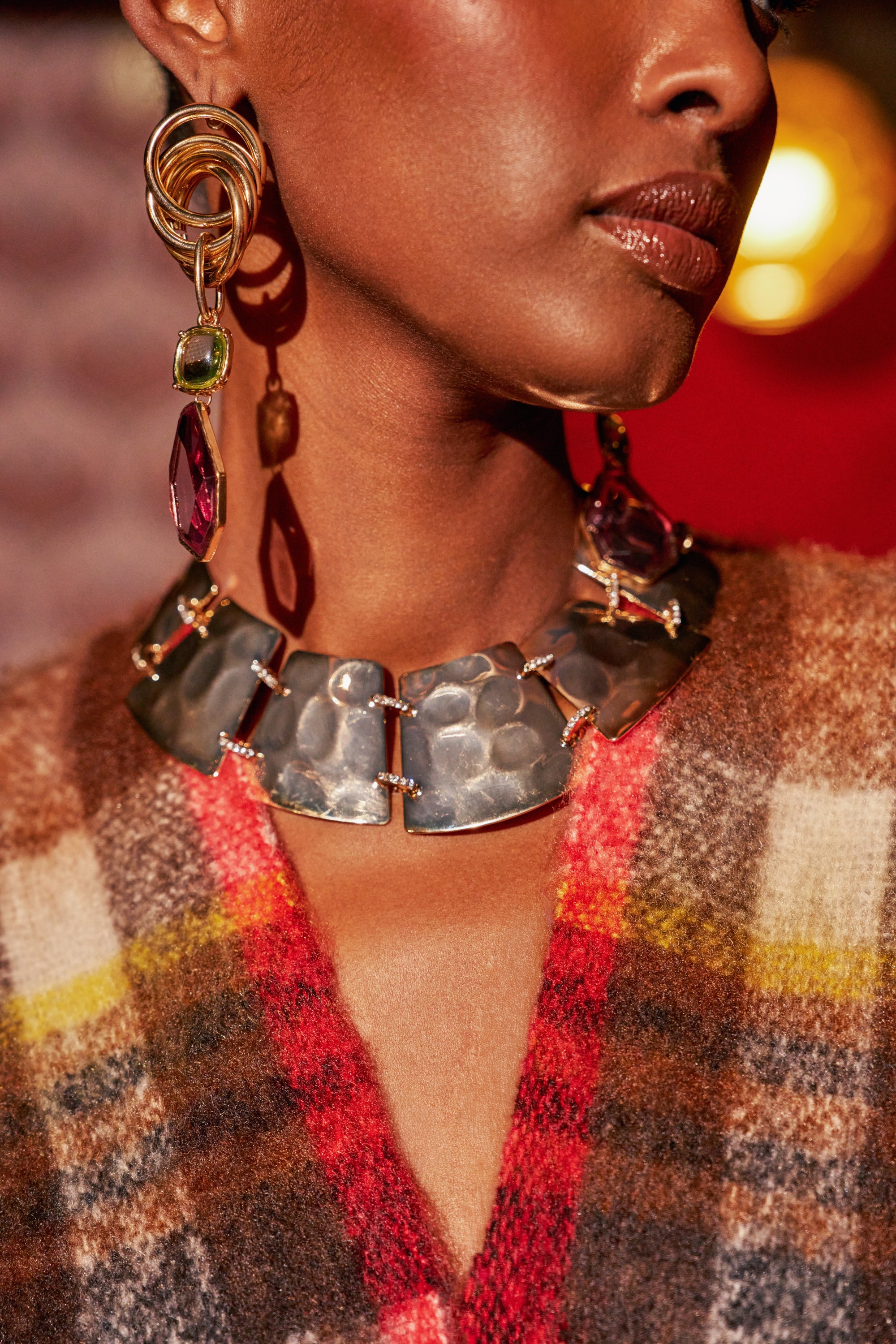 Close-up of a model wearing a GILDED ARMOUR NECKLACE, which is a wide, hammered gold collar necklace, paired with long, multi-stone drop earrings.