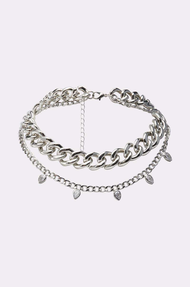The TOO RICH NECKLACE is a silver-tone layered statement piece with thick and thin chains, adorned with small teardrop charms on the delicate chain, shown on a plain light background.