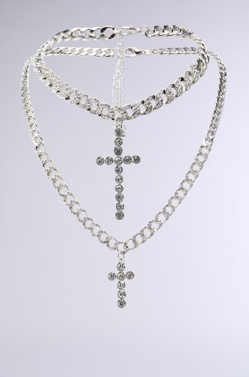 The ICED OUT NECKLACE SET features a multi-layered silver rhinestone design with two detailed cross pendants, each adorned with clear gemstones, hanging from varying lengths of chain links, and is presented against a plain light grey background. 1