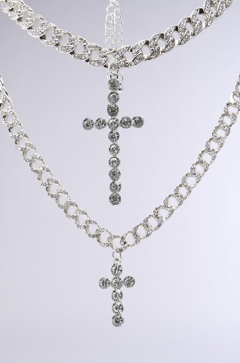 The ICED OUT NECKLACE SET features two silver Cuban chain necklaces with cross pendants, beautifully displayed against a light grey background. Each cross is encrusted with small, sparkling gemstones, and the chains boast a thick, interlocking link design. The rhinestone necklaces are perfectly layered, with one hanging slightly lower than the other. 2