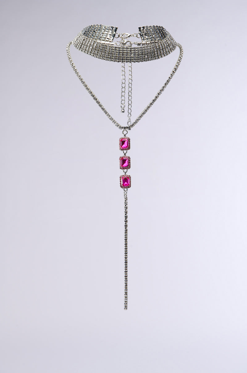 Introducing the NIKKI LONG NECKLACE: a luxury piece featuring a silver choker with an intricate grid design, complemented by a long chain pendant adorned with three large rectangular pink gemstones. The statement accessory's lower portion includes a dangling silver chain, all set against a plain gray background.
