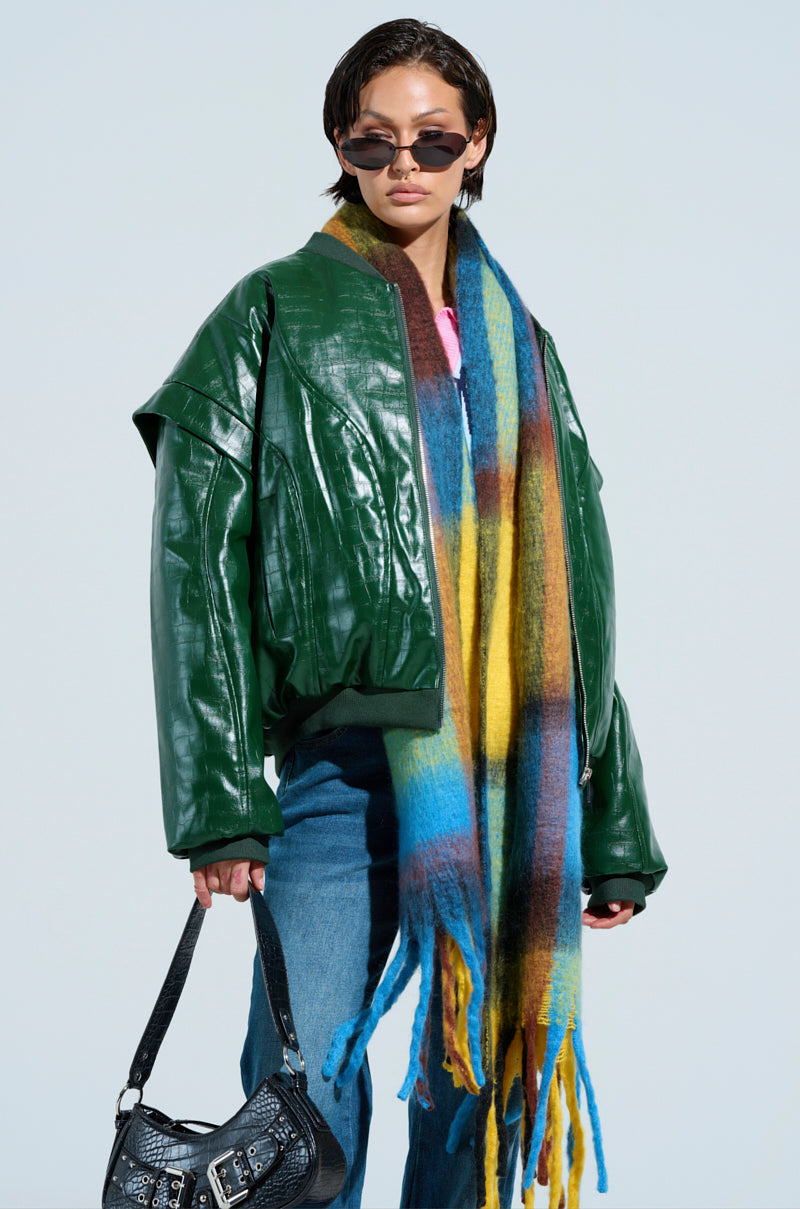 A person wearing a dark green YOKON embossed faux leather bomber, colorful scarf, and sunglasses holds a black handbag. They are dressed in blue jeans against a plain background.
