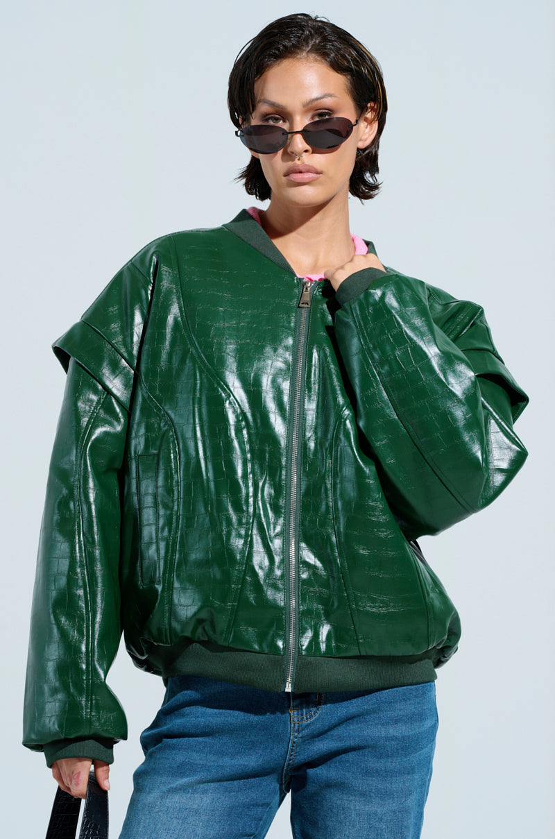 Sporting blue jeans and dark sunglasses, a person poses confidently against a light blue backdrop. Their short hair complements the YOKON EMBOSSED FAUX LEATHER BOMBER with its vegan crocodile finish. They hold a black bag in one hand.