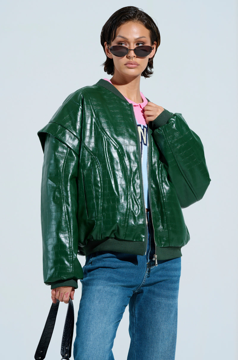 A person with short hair is wearing the YOKON EMBOSSED FAUX LEATHER BOMBER, a shiny green jacket with a crocodile pattern, over a pink shirt. They're in blue jeans and sunglasses, holding a black accessory against a light blue background.