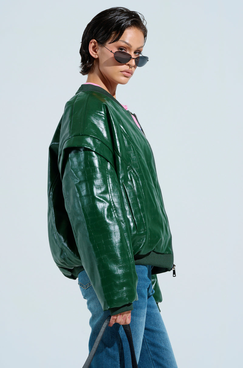 Wearing the YOKON EMBOSSED FAUX LEATHER BOMBER in shiny green with an embossed crocodile pattern, a person poses confidently in sunglasses against a light background. They pair it with blue jeans and hold a bag, exuding a stylish and relaxed vibe.