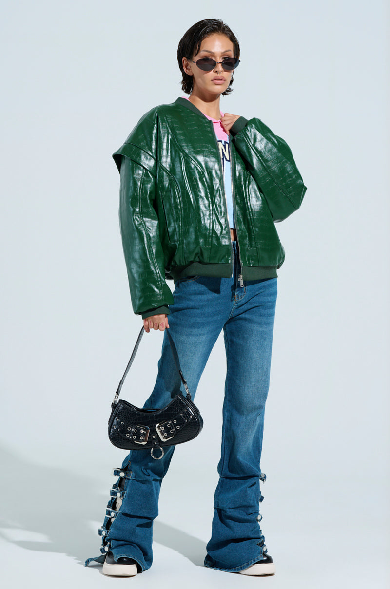 A person with short hair wears a glossy green YOKON EMBOSSED FAUX LEATHER BOMBER, blue flared jeans with cutouts and embellishments, white sneakers, and sunglasses while holding a black handbag. The plain background highlights the vegan faux leather fashion statement.
