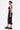 A woman in profile wears the ONE OF ONE ABSTRACT MIDI DRESS with a side cutout, large hoops, a chunky chain necklace, and black platform sandals with gold studs. Her neat low bun completes her avant-garde look.