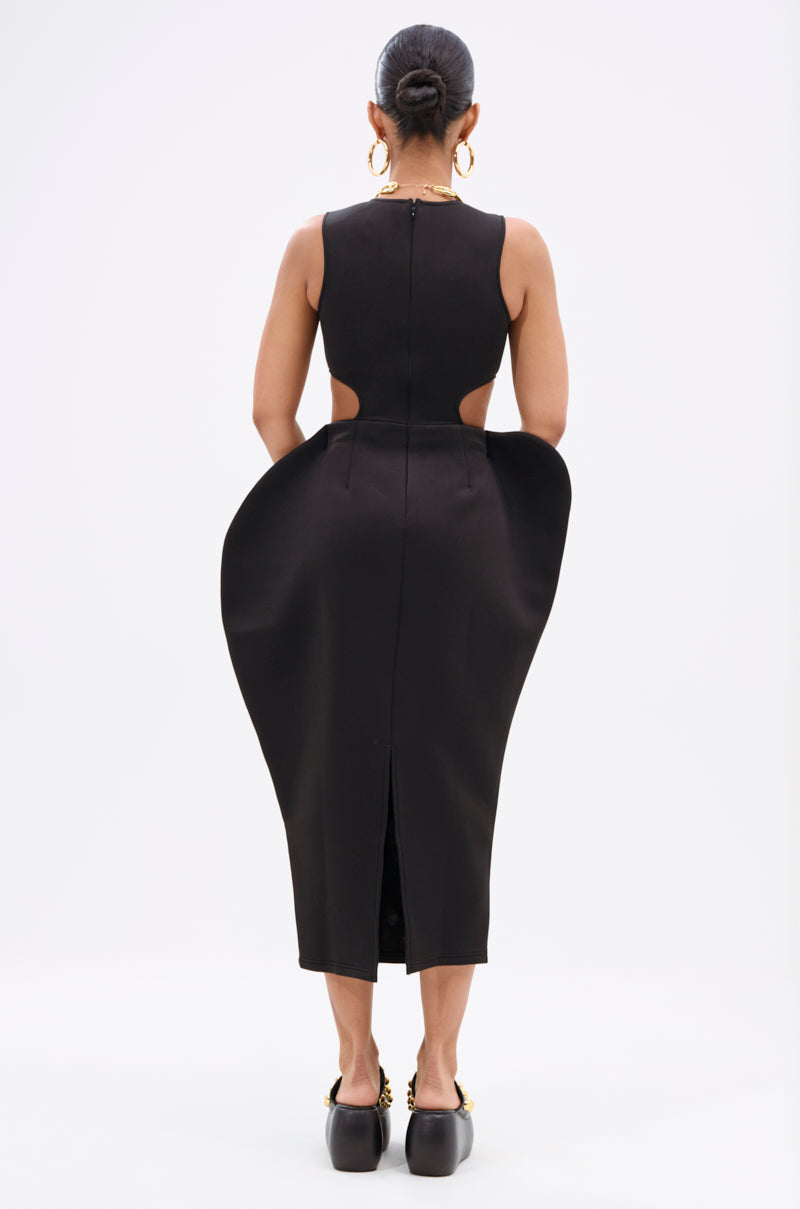 A woman stands facing away, wearing the ONE OF ONE ABSTRACT MIDI DRESS with sculpted hips, side cutouts, and a mid-calf hem. Her hair is in a tight bun, paired with gold hoop earrings and black platform shoes.
