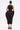 A woman stands facing away, wearing the ONE OF ONE ABSTRACT MIDI DRESS with sculpted hips, side cutouts, and a mid-calf hem. Her hair is in a tight bun, paired with gold hoop earrings and black platform shoes.