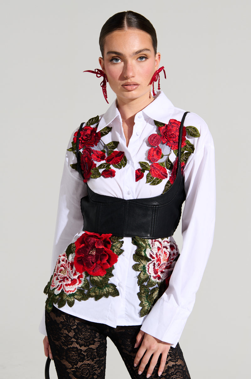 A person wearing the ROSA CORSET DETAIL BUTTON DOWN BLOUSE, featuring red and green floral appliques beneath a black waist cincher, accessorizes with red statement earrings and black lace leggings. Their hair is slicked back as they pose with one hand holding a black strap.
