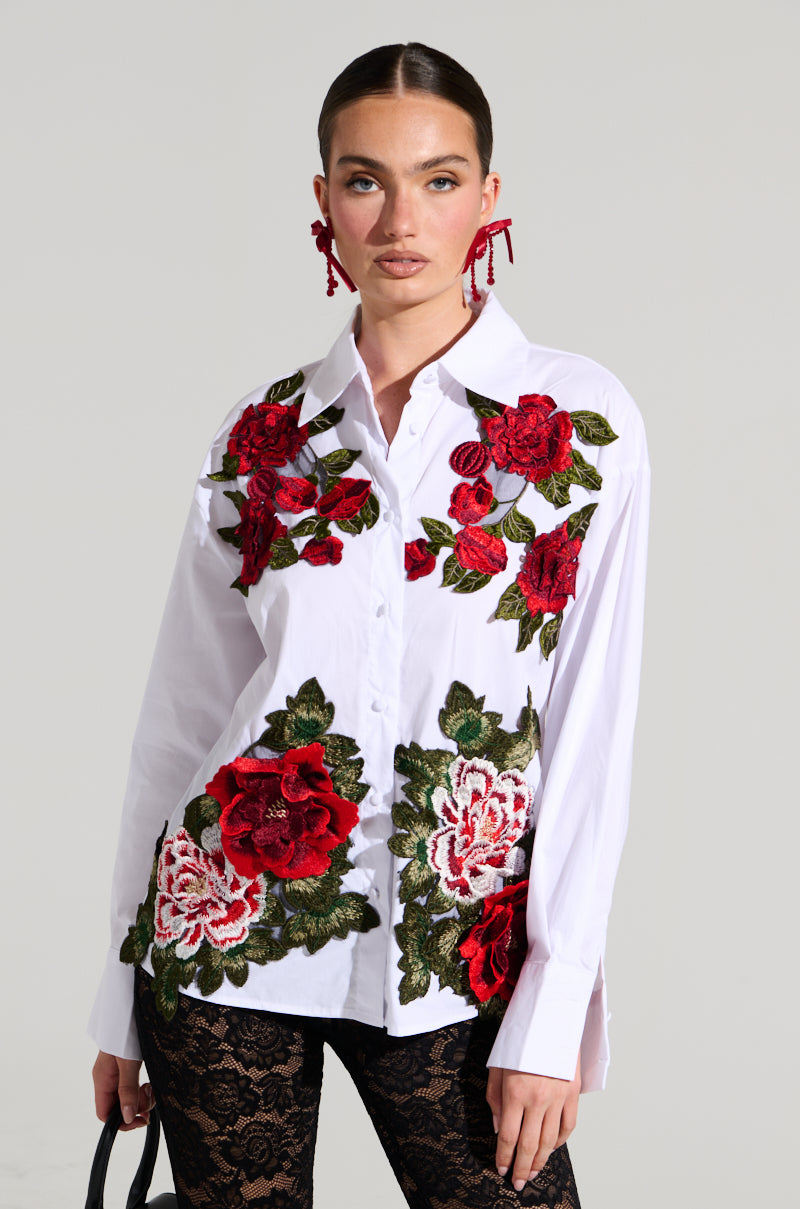 A woman with a serious expression is wearing the ROSA CORSET DETAIL BUTTON DOWN BLOUSE, featuring large red and white floral embroidery on the front. She also wears black lace pants and red floral earrings, holding a black handbag by her side. The background is plain light grey.