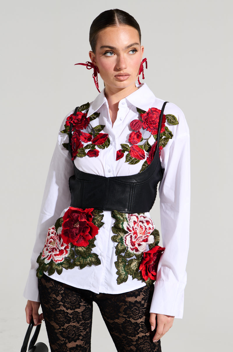 A person wearing the ROSA Corset Detail Button Down Blouse, featuring white fabric with red and pink floral embroidery, paired with a black corset with intricate details. They also wear black lace leggings and accessorize with red ribbon earrings while holding a small black handbag. The background is a plain light gray.
