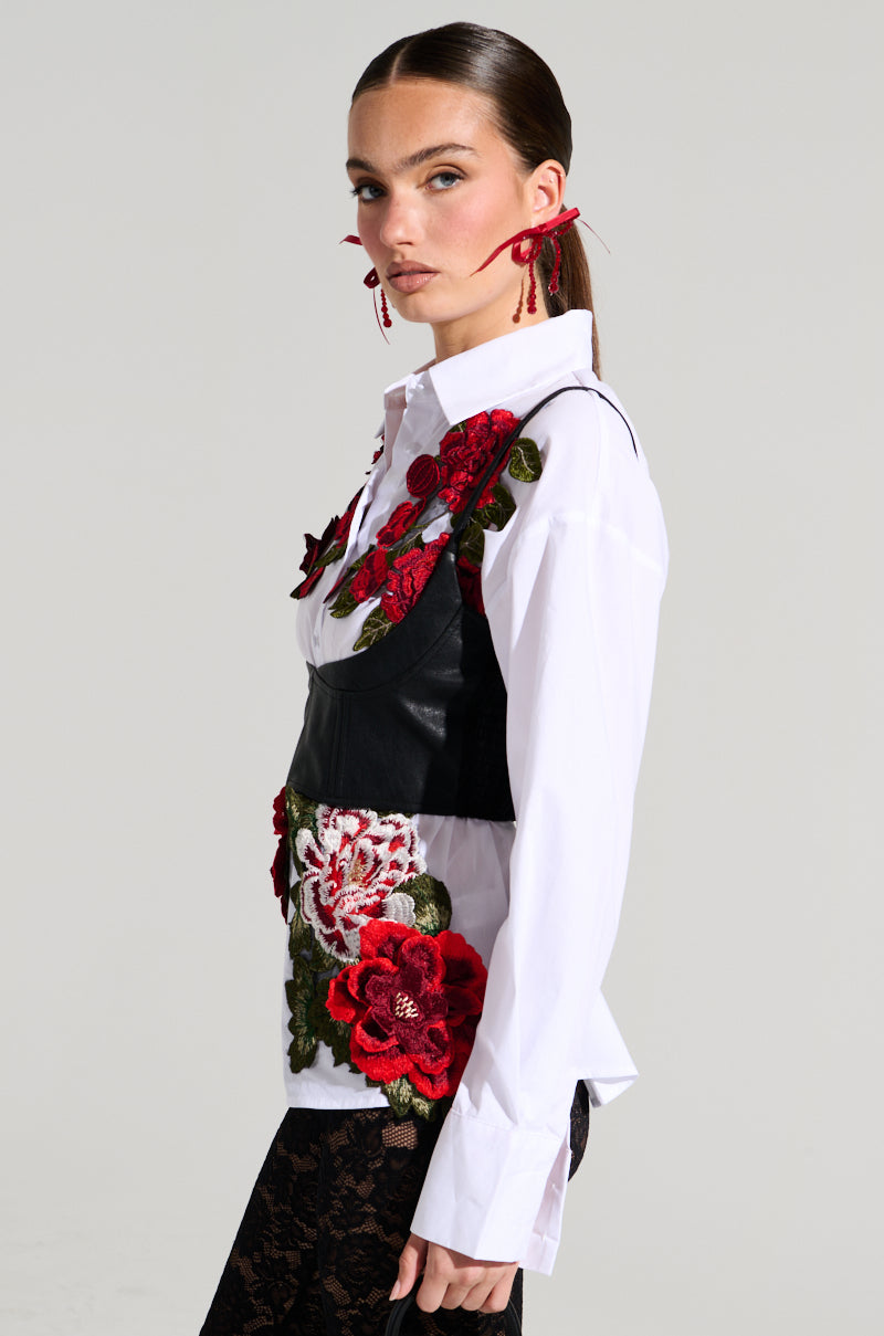 A woman with her hair pulled back, wearing the ROSA CORSET DETAIL BUTTON DOWN BLOUSE adorned with intricate red and green floral appliques, stands against a plain background. She has red earrings and black lace pants. She gazes confidently towards the camera.