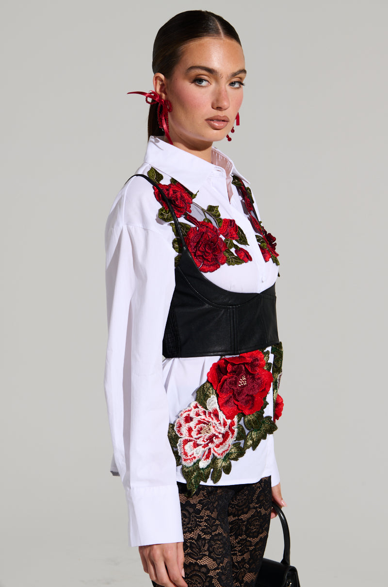 A woman with sleek hair tied back and red ribbon earrings wears the ROSA Corset Detail Button Down Blouse, which is adorned with vibrant red floral appliques. She completes her outfit with a black bustier featuring corset details layered over the blouse, black lace-patterned pants, and is holding a small black bag.