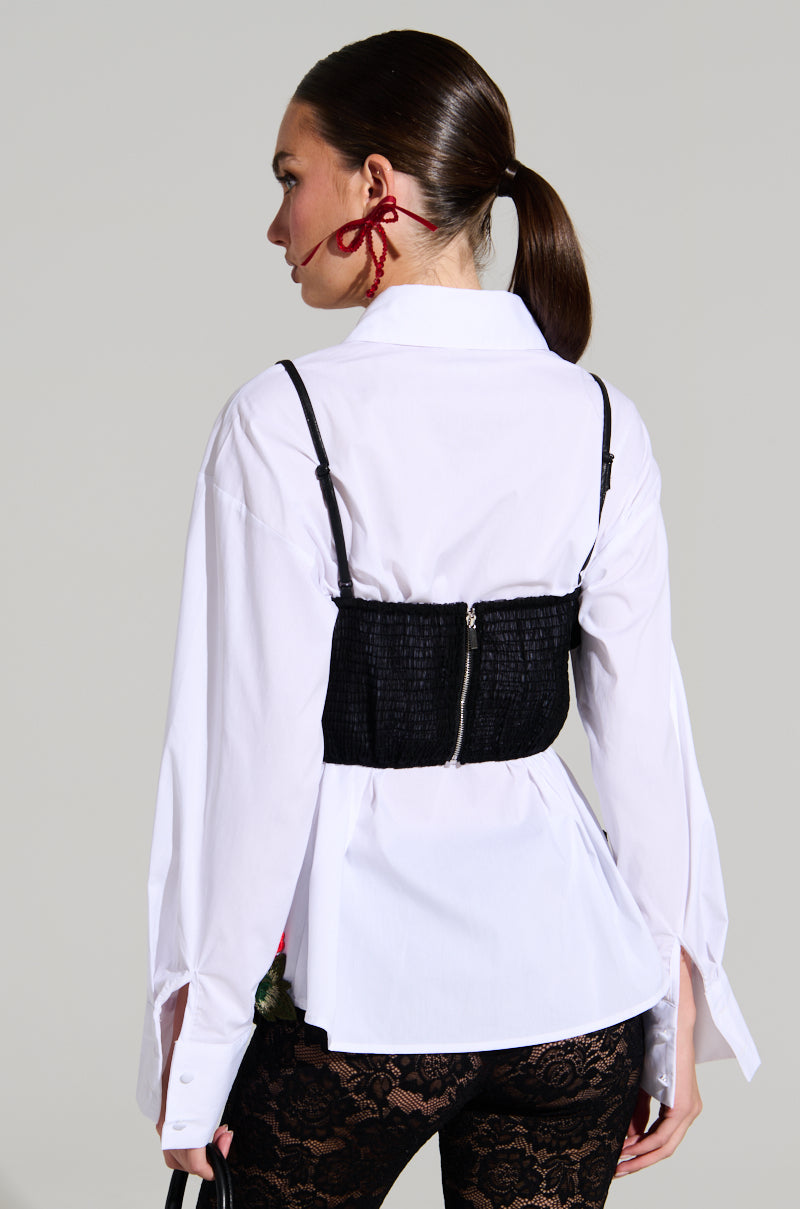 A person with long, dark hair tied in a ponytail is shown from behind, wearing the ROSA CORSET DETAIL BUTTON DOWN BLOUSE paired with black lace pants. Bold red earrings dangle, and their right hand rests on their waist, highlighting the corset detail of the blouse.