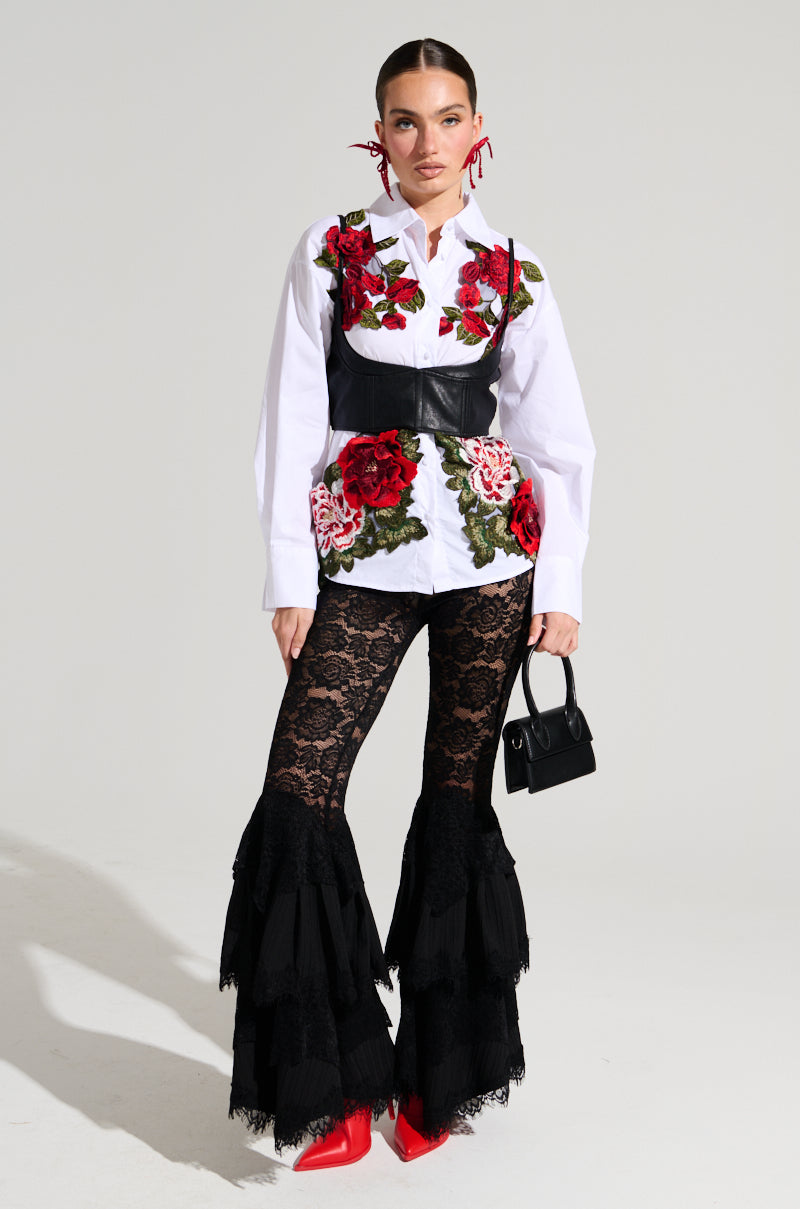 A woman stands against a plain background wearing the ROSA CORSET DETAIL BUTTON DOWN BLOUSE, a white top adorned with red and pink floral appliques. She pairs it with black lace pants featuring layered ruffles at the bottom, red pointed shoes, and a small black handbag. Her hair is tied back.