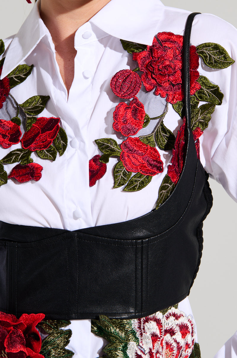 Close-up of a person wearing the ROSA CORSET DETAIL BUTTON DOWN BLOUSE featuring a white base with elaborate red and green floral appliques. Only the torso and part of the neck are visible in the image, emphasizing the intricate corset-like detailing.