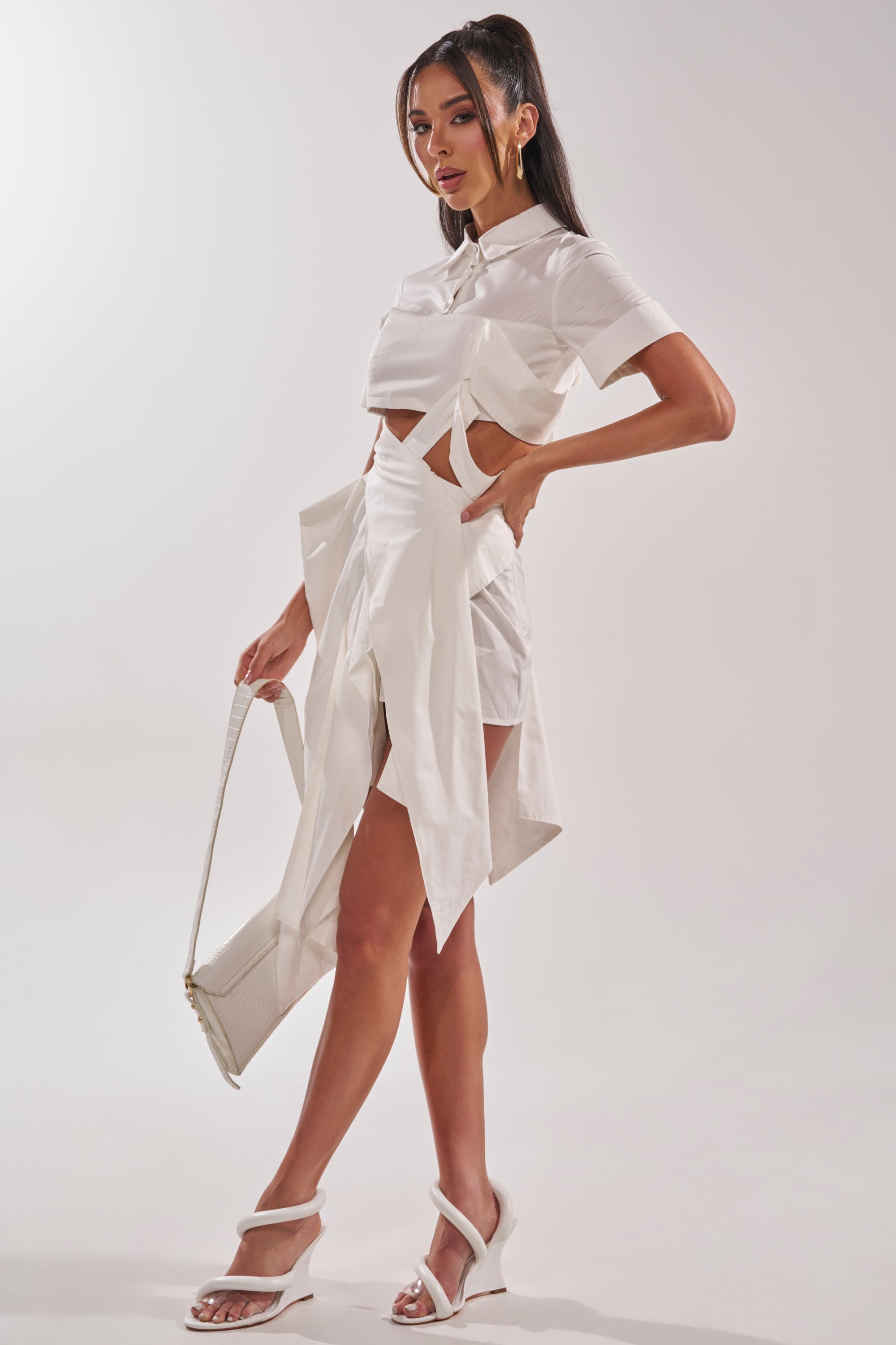 A woman poses in the modern white RIRI CUT OUT POPLIN MIDI DRESS, paired with white heeled sandals and a white handbag, against a plain light background.