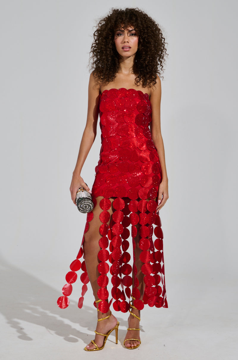 A woman with curly hair is wearing the SHE IS THE MOMENT RHINESTONE MAXI DRESS, showcasing a strapless neckline. She complements the look with gold high-heeled sandals and holds an ornate purse, her ensemble exuding sophistication and style.