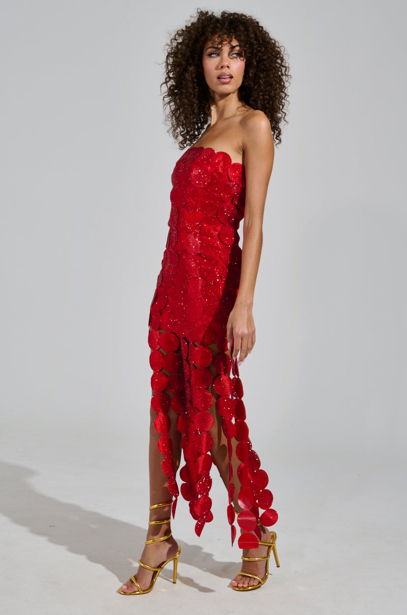 A person with curly hair wears the SHE IS THE MOMENT rhinestone maxi dress featuring a strapless neckline, paired elegantly with gold strappy high heels, set against a plain gray backdrop.