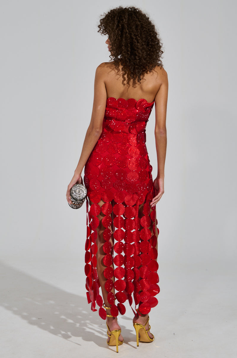 The woman with curly hair is wearing the SHE IS THE MOMENT RHINESTONE MAXI DRESS, characterized by its strapless red design featuring large circular sequins and a fringed skirt. She completes her elegant look with a small silver clutch and stands in yellow high heels against a gray background, emphasizing the dress's sophisticated strapless neckline.