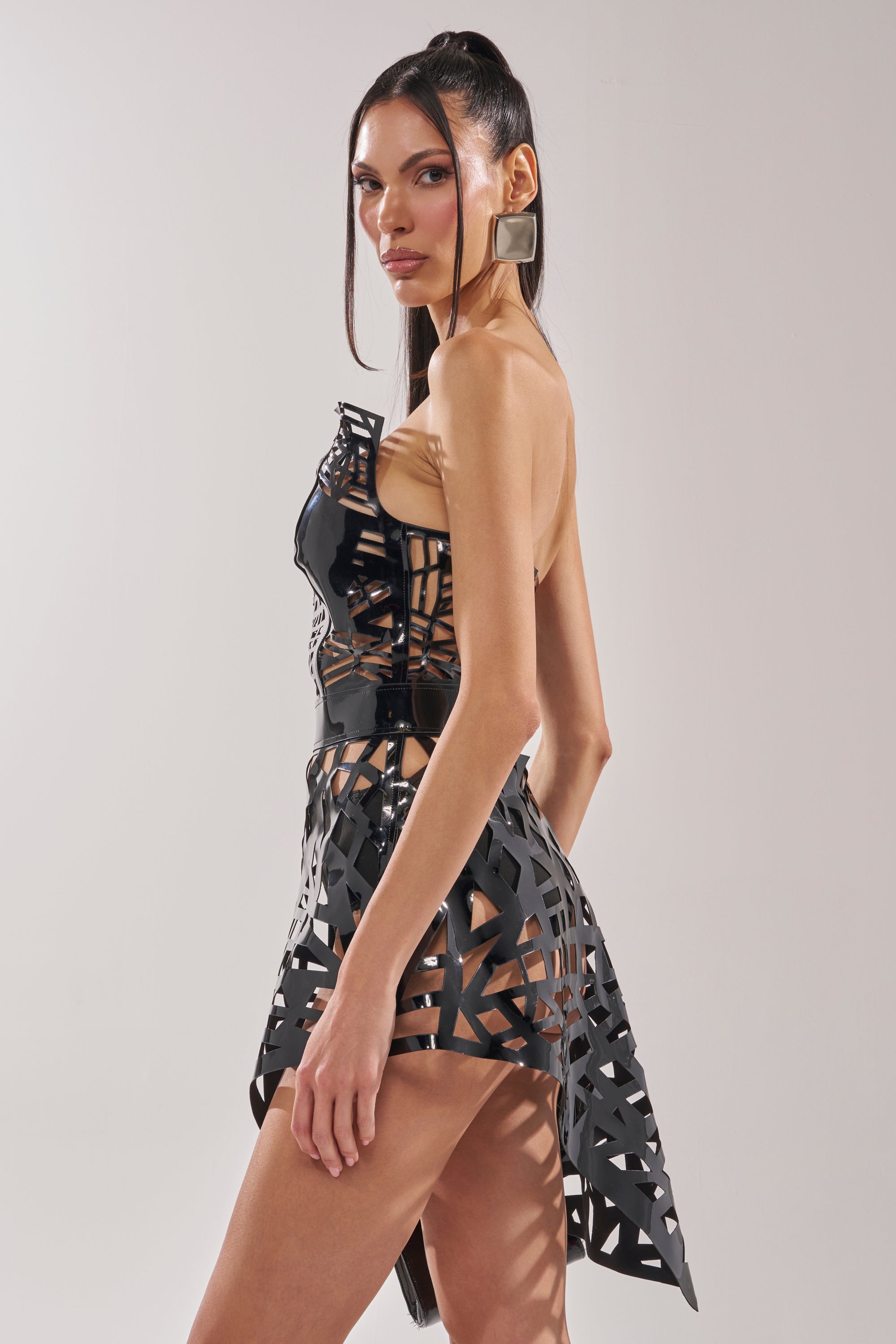 A woman poses sideways in the PARTY IN THE BACK CUT OUT MINI DRESS—black with sheer sections—paired with large square earrings and a high ponytail, set against a plain light background.