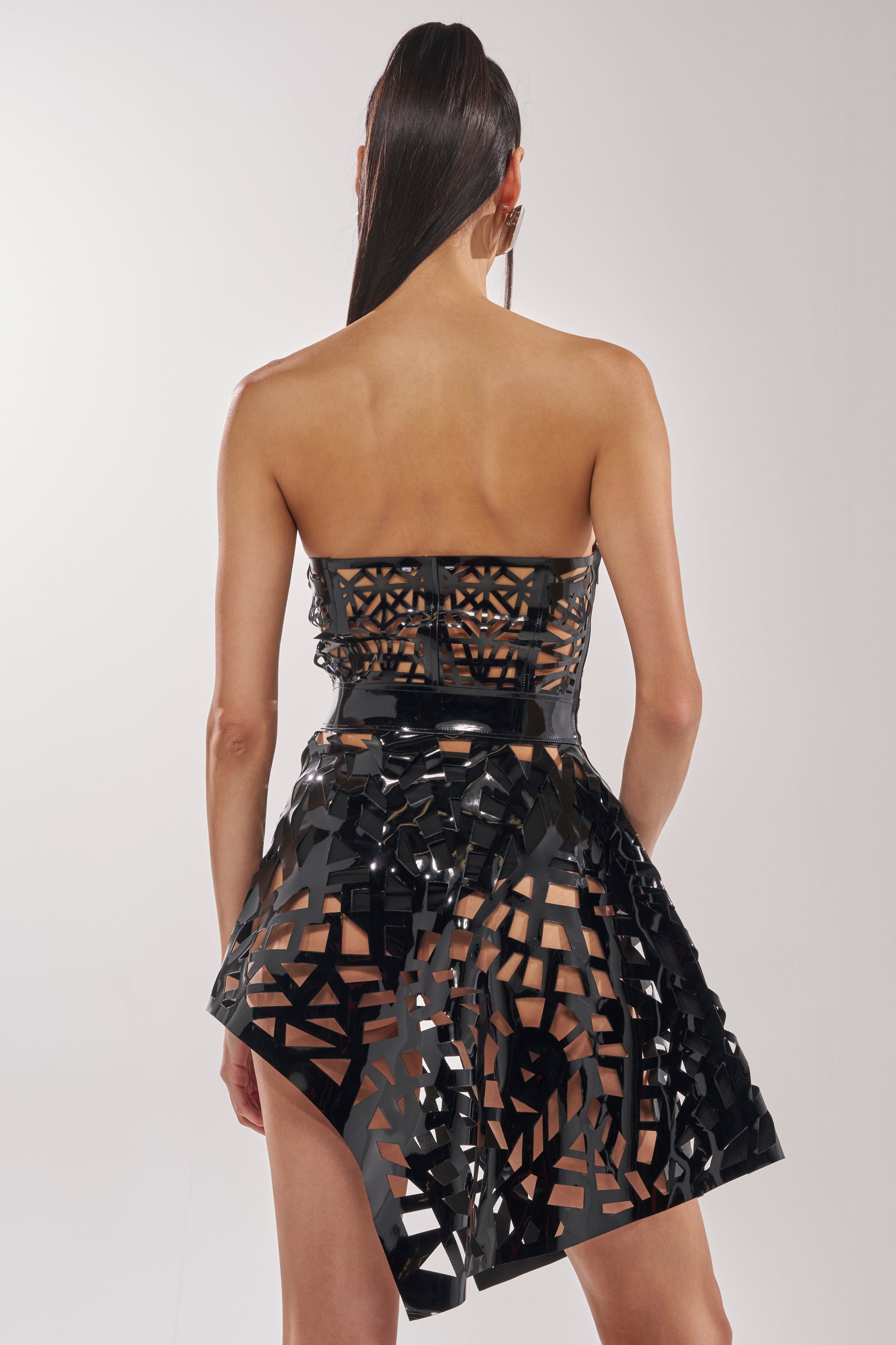 A woman with long, straight hair tied back is seen from behind wearing the PARTY IN THE BACK CUT OUT MINI DRESS—a shiny black strapless mini dress with geometric cutouts and an asymmetrical skirt. The background is plain and light-colored.