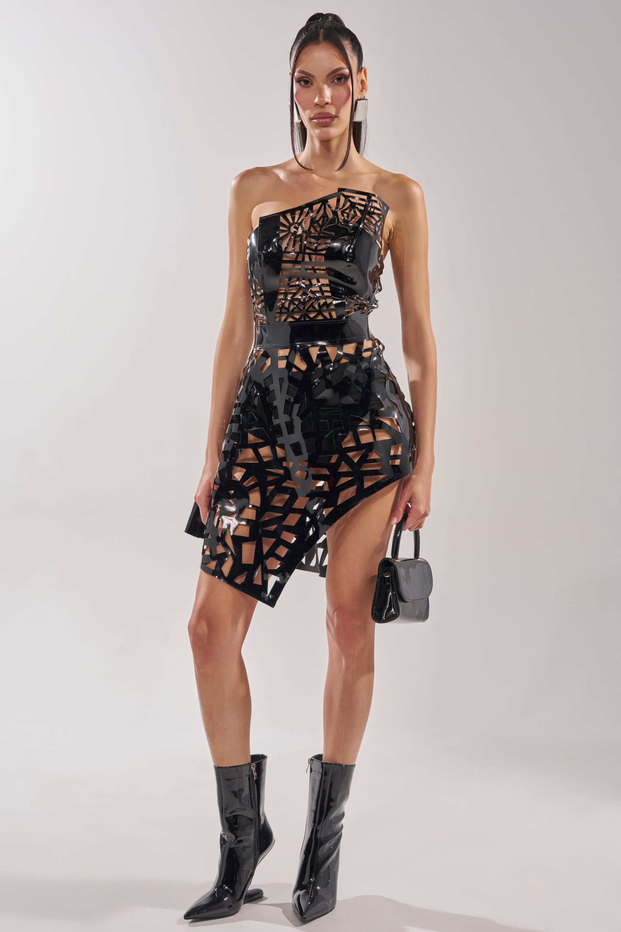 A woman poses in the PARTY IN THE BACK CUT OUT MINI DRESS, featuring a shiny, asymmetrical cut-out design with geometric details, styled with matching black boots and a small black handbag against a plain light background.