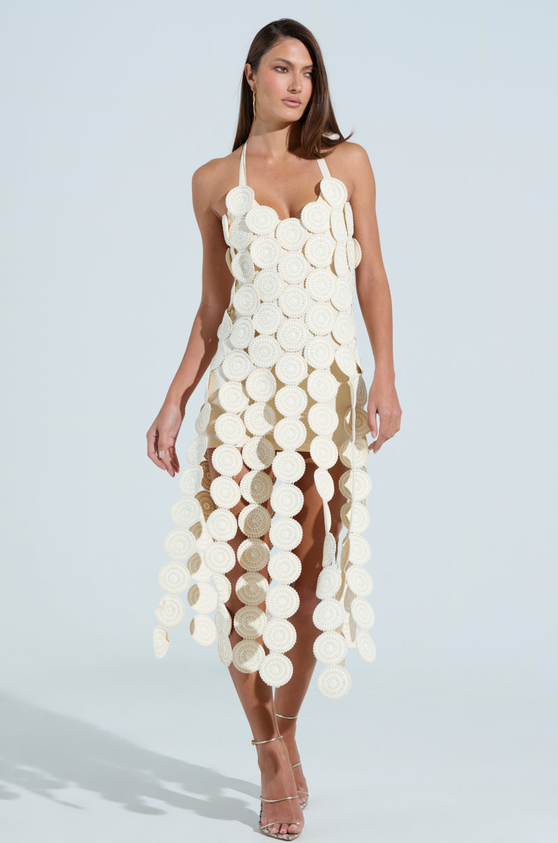 A woman poses confidently in an IBIZA DREAMIN TEXTURED MIDI DRESS with large white circular discs and cutouts. The sleeveless, ankle-length dress has a chic tie-back halter neck. She pairs it with strappy heels against a plain light background.