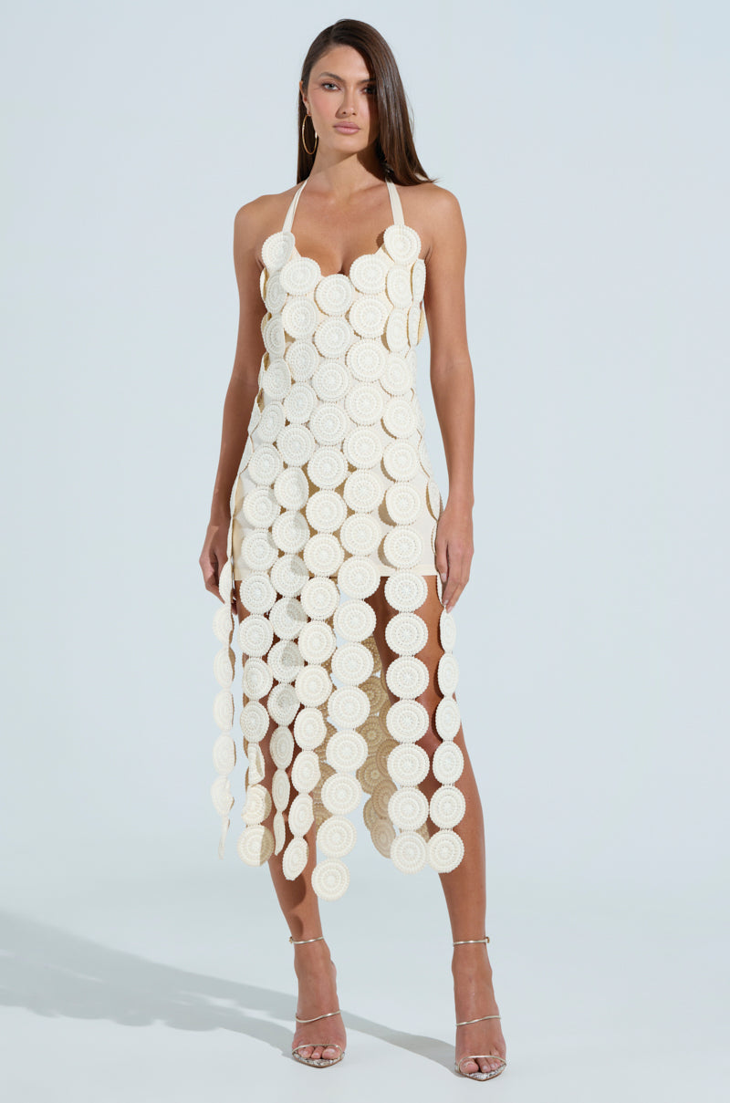 A woman poses in the Ibiza Dreamin Textured Midi Dress, a mid-length halter-neck design with circular cream discs and a fringe hem. She complements it with silver high heels and hoop earrings, her straight shoulder-length hair against a light blue backdrop.