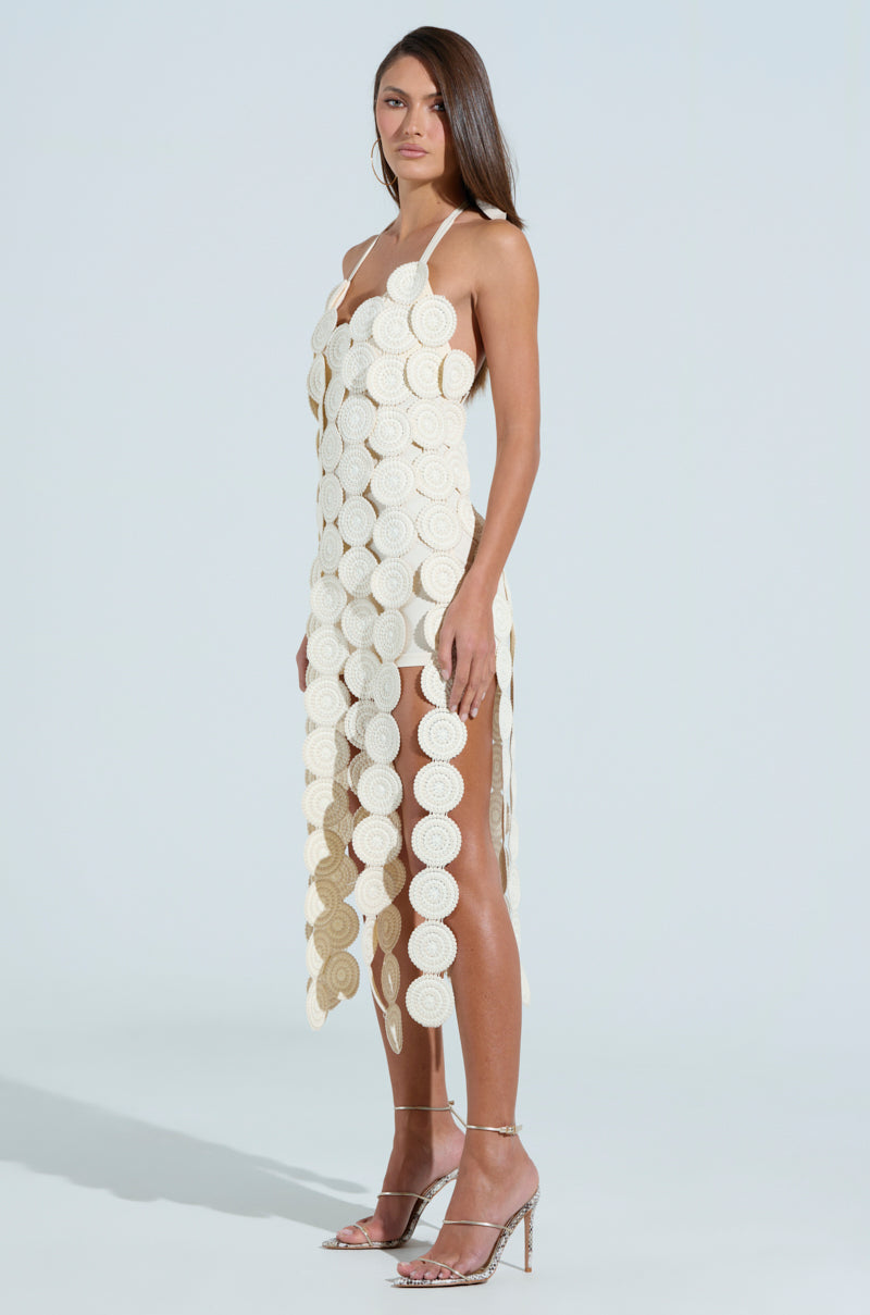 A woman poses against a light blue background in the IBIZA DREAMIN TEXTURED MIDI DRESS, featuring circular off-white discs and a chic halter neck. Clear high-heeled shoes complete her elegant, effortlessly charming look.