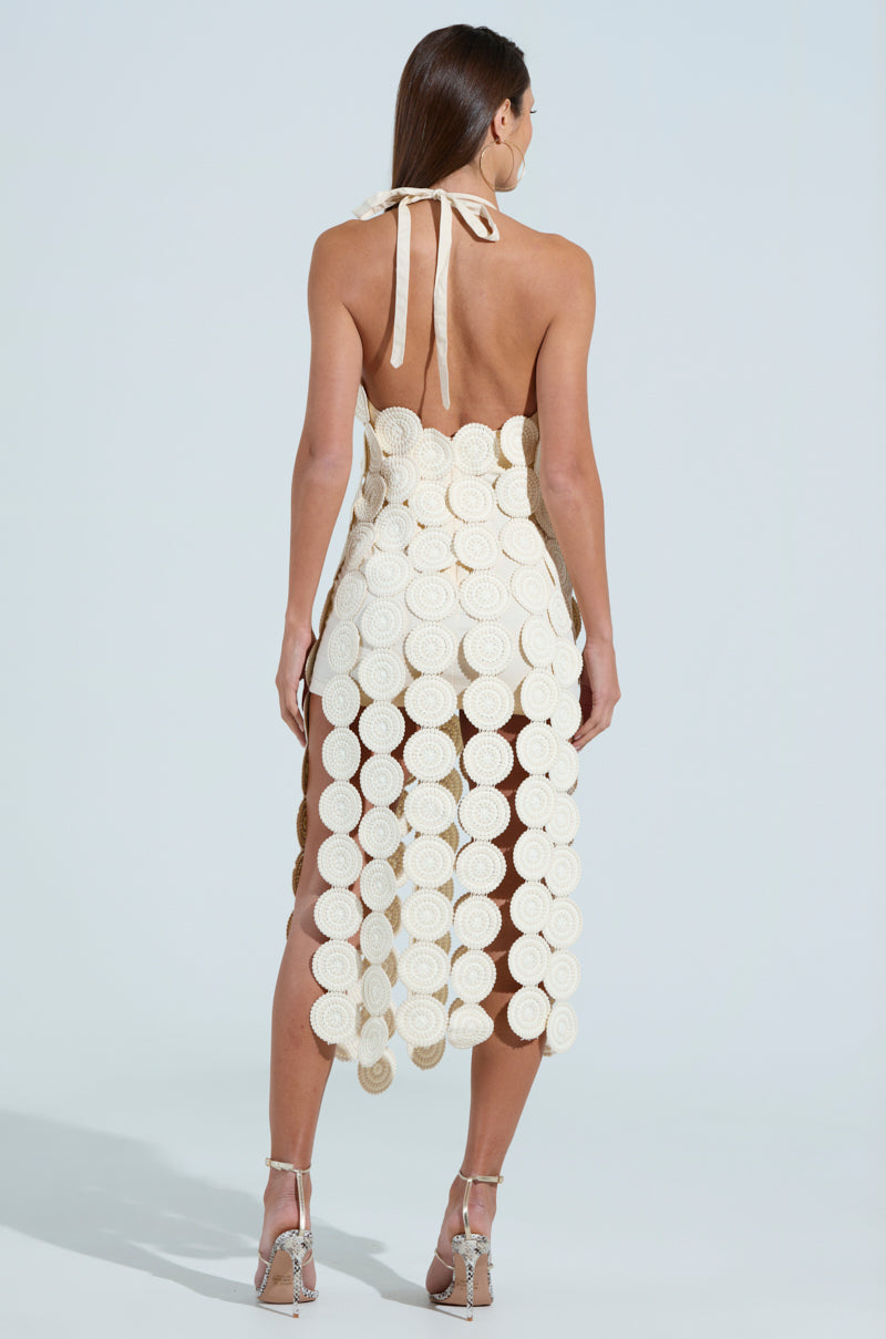 A woman poses with her back to the camera, wearing the backless IBIZA DREAMIN TEXTURED MIDI DRESS featuring circular patterns. The ankle-length design is elegantly paired with high heels, and her long hair flows against a plain, light-colored backdrop.