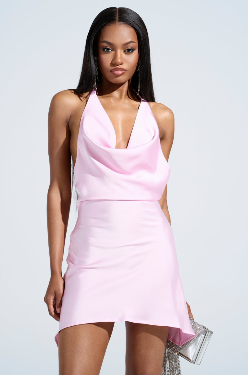 A woman stands confidently in the DOLLY SATIN MINI DRESS IN LIGHT PINK, featuring a satin finish with a cinched waist and halter neckline. Her long, straight dark hair complements her silver clutch against the plain, light-colored backdrop, highlighting her elegant ensemble.