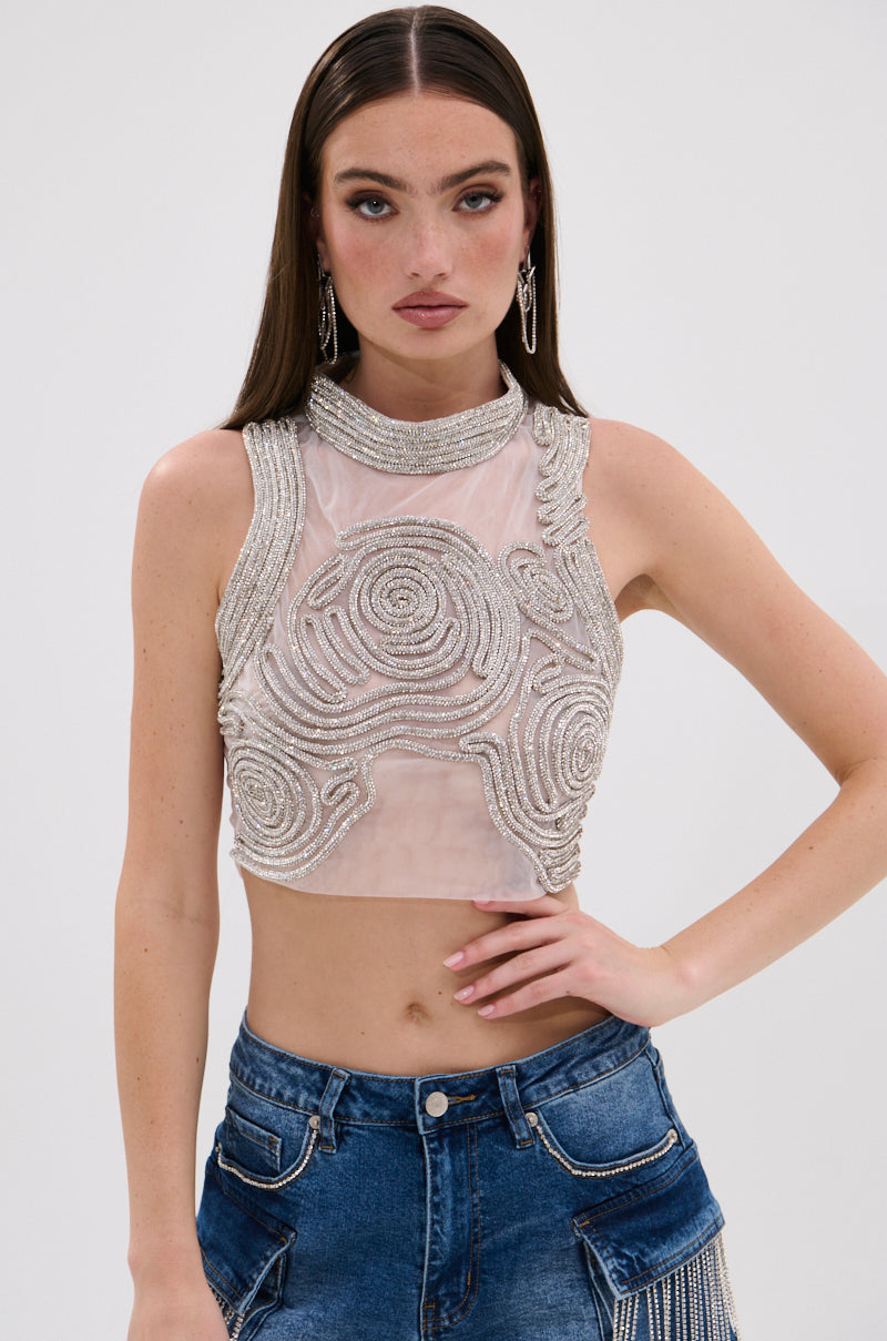 A woman with long brown hair wears the STARSHINE RHINESTONE MOCK NECK BLOUSE, a sheer mesh tank adorned with silver bead swirls, paired with high-waisted blue jeans featuring decorative stitching, against a plain white background. 0