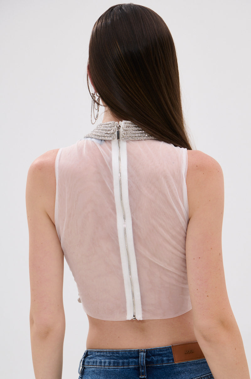 A woman with long brown hair, seen from behind, wears the STARSHINE RHINESTONE MOCK NECK BLOUSE—a sleeveless sheer white crop top with an embellished collar and visible back zipper—paired with blue jeans. 4
