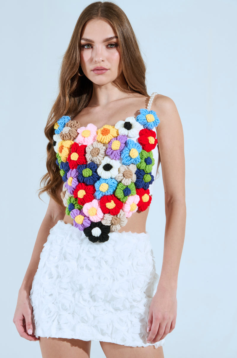 A woman with long hair stands against a light blue background, wearing the vibrant PENELOPE FLORAL CORSET TOP paired with a white skirt featuring a textured floral pattern adorned with delicate pearl beads.