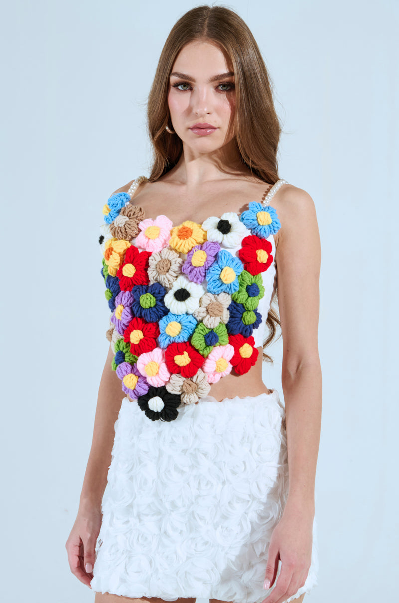 A woman with long brown hair wears a Penelope Floral Corset Top with colorful crocheted flowers and pearl beads, paired with a floral-patterned white skirt. She stands against a plain background.