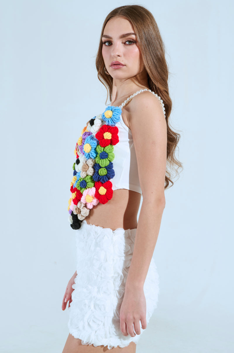 A person with long brown hair is wearing a PENELOPE FLORAL CORSET TOP adorned with colorful pom-poms and pearl beads, paired with a white textured skirt. They stand in profile against a light blue background.