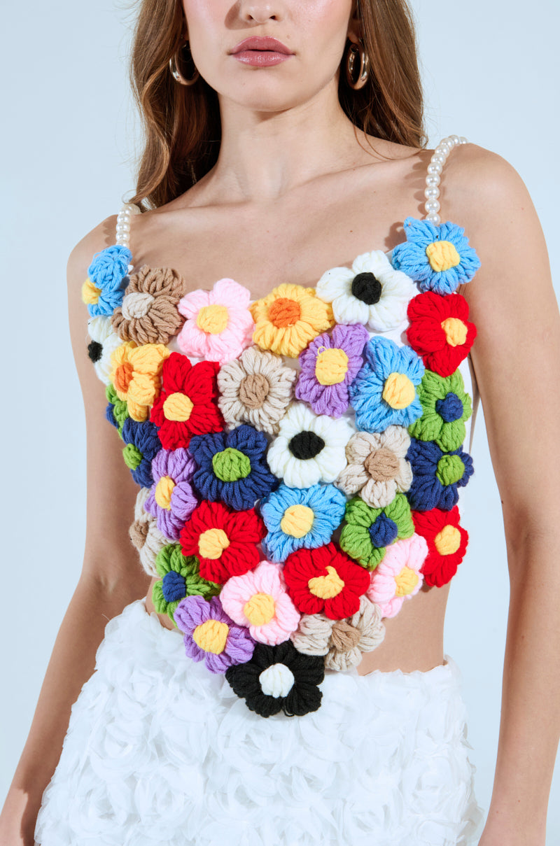 A woman wears the PENELOPE FLORAL CORSET TOP, a vibrant floral crocheted piece with colorful yarn flowers and pearl beads, paired with a white textured skirt against a plain, light-colored background.