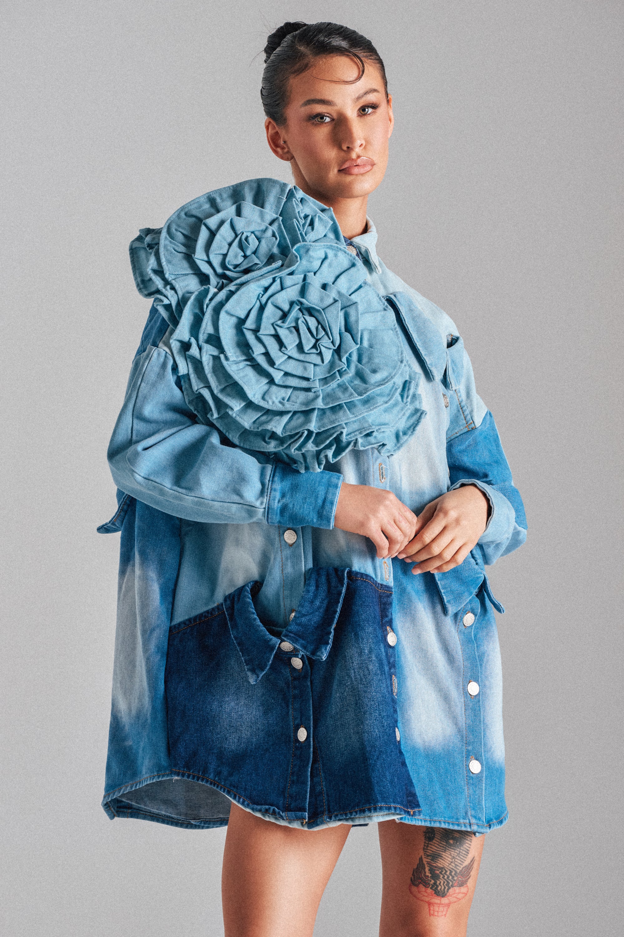 A woman with her hair tied back and a visible thigh tattoo wears the SO FINE DENIM BROOCH shirt, featuring oversized sculpted floral designs on the shoulder, while standing against a plain gray background.