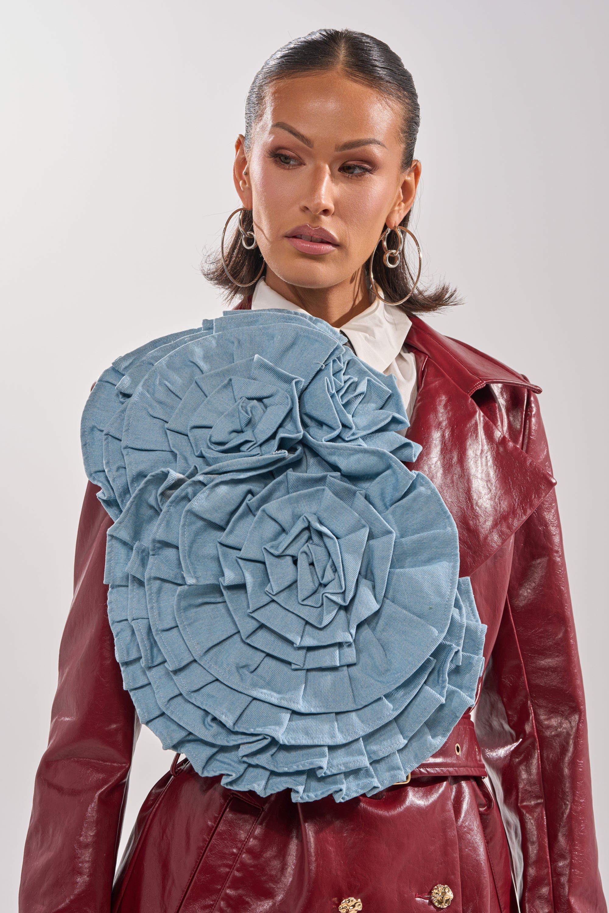 A woman with slicked-back hair wears a shiny maroon trench coat, a SO FINE DENIM BROOCH on her shoulder, large hoop earrings, and a white collared shirt. 0