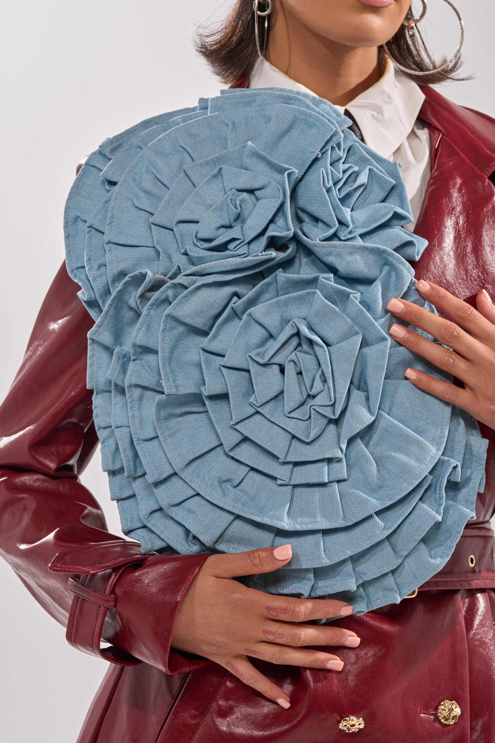 A woman in a red leather jacket and white shirt holds the SO FINE DENIM BROOCH, a large, textured blue clutch shaped like layered spiral flowers. Only her lower face, hands, and the bag are visible.