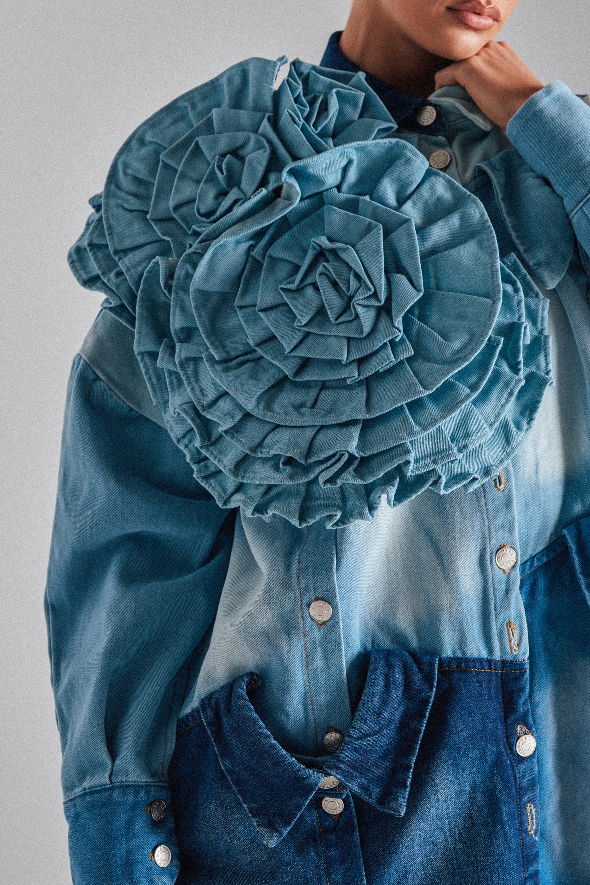 Wearing a sky blue denim jacket accented with the SO FINE DENIM BROOCH on the shoulder and chest, a person poses with one hand near their chin.