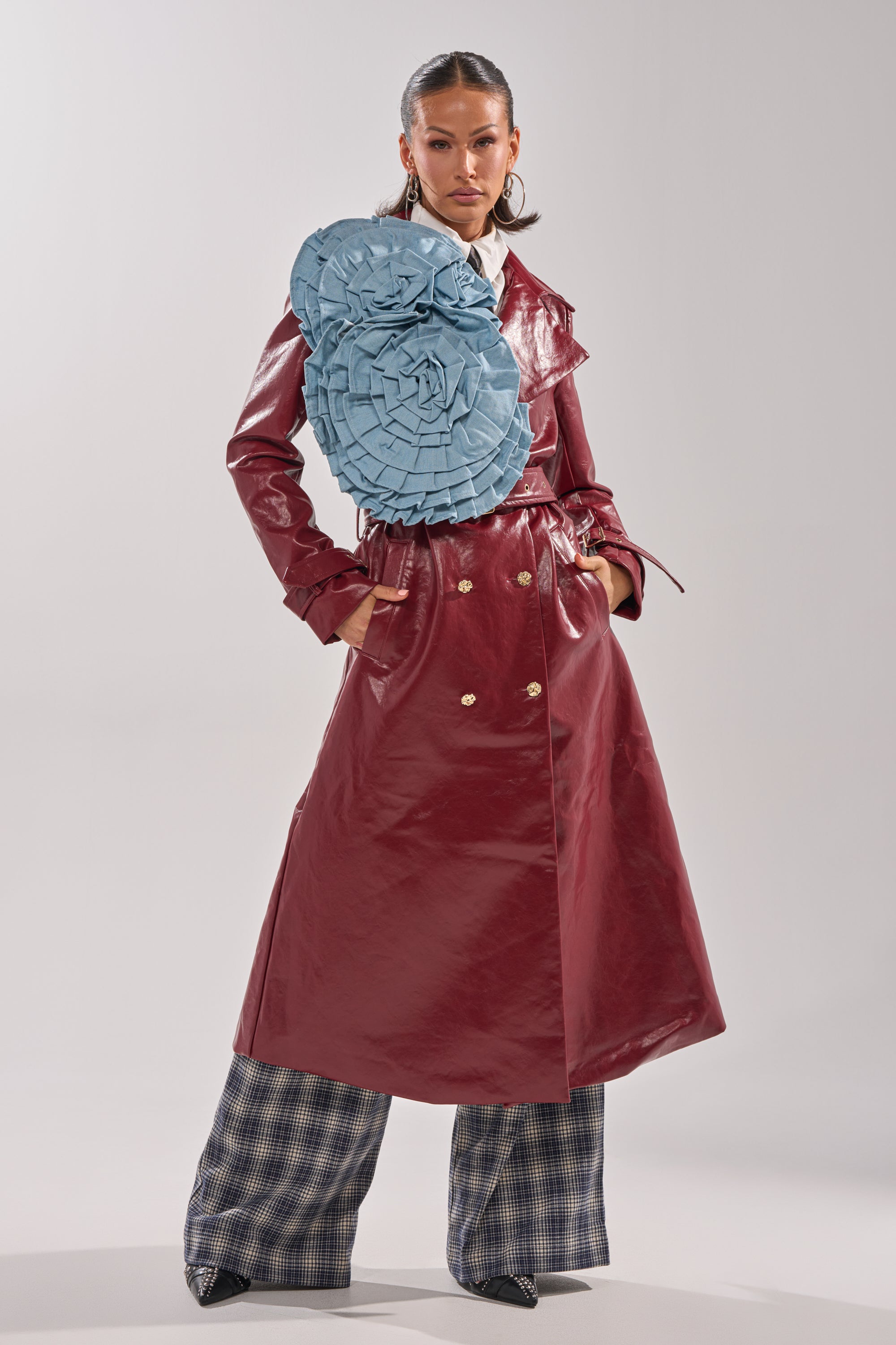 Wearing a shiny maroon trench coat, wide plaid pants, pointed shoes, and the SO FINE DENIM BROOCH on one shoulder, a person stands confidently against a plain light background.