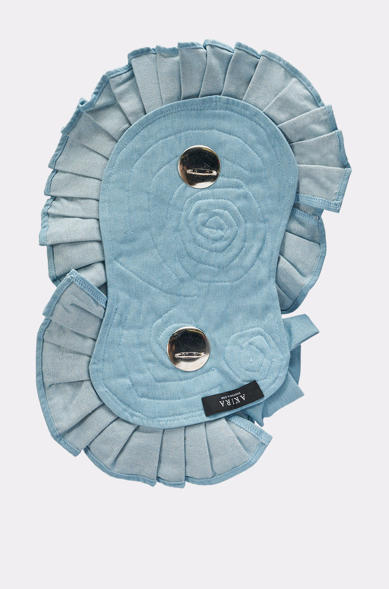 The SO FINE DENIM BROOCH features light blue quilted fabric with pleated ruffle edges, two round metallic buttons, an embroidered floral design resembling a delicate flower, and a visible "AXIA" label near the bottom center.