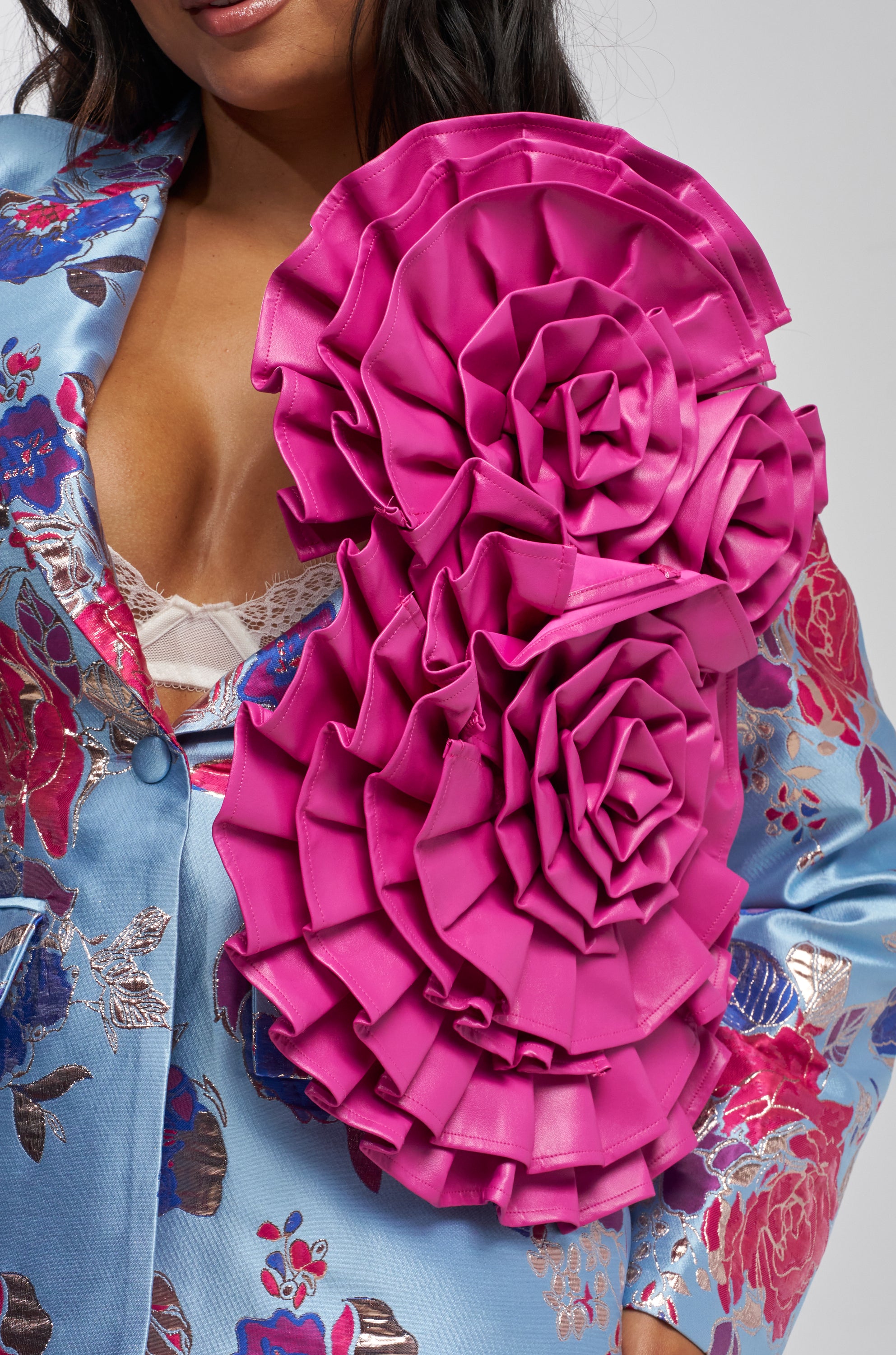 A person in a light blue floral blazer decorated with IN MY POWER PU STATEMENT BROOCHES—large, layered pink faux leather flowers on the shoulder and chest—partially reveals a lacy white bra underneath.