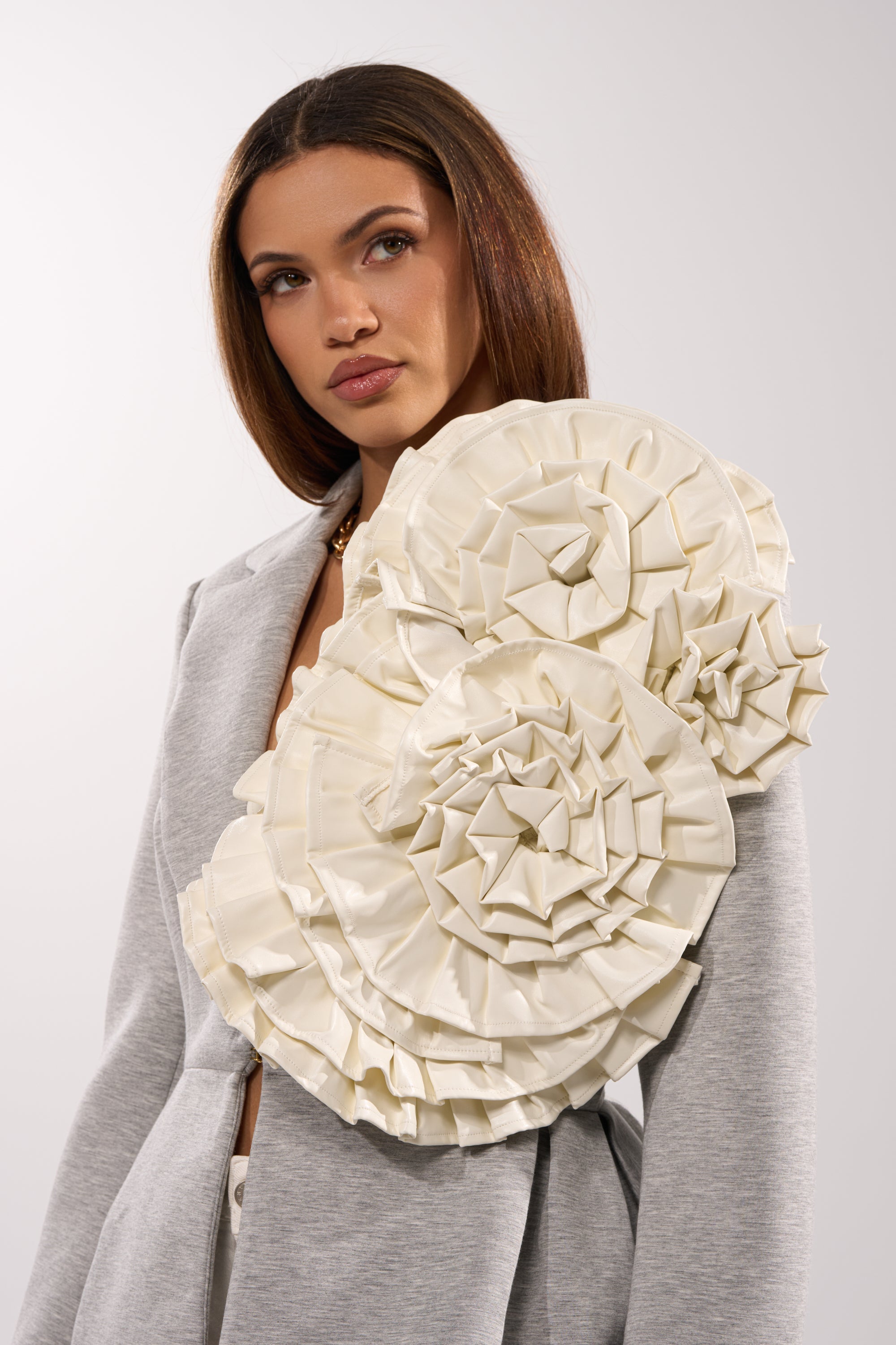A woman with straight brown hair wears a light gray blazer adorned with the BLOOMING PU OVERSIZED BROOCH—an eye-catching sculptural white floral accessory on one shoulder—against a plain light background. 0