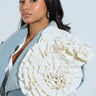 A woman with long dark hair wears the BLOOMING PU OVERSIZED BROOCH on a gray jacket adorned with an intricate white floral applique on the shoulder, standing against a simple light background, gazing calmly at the camera.