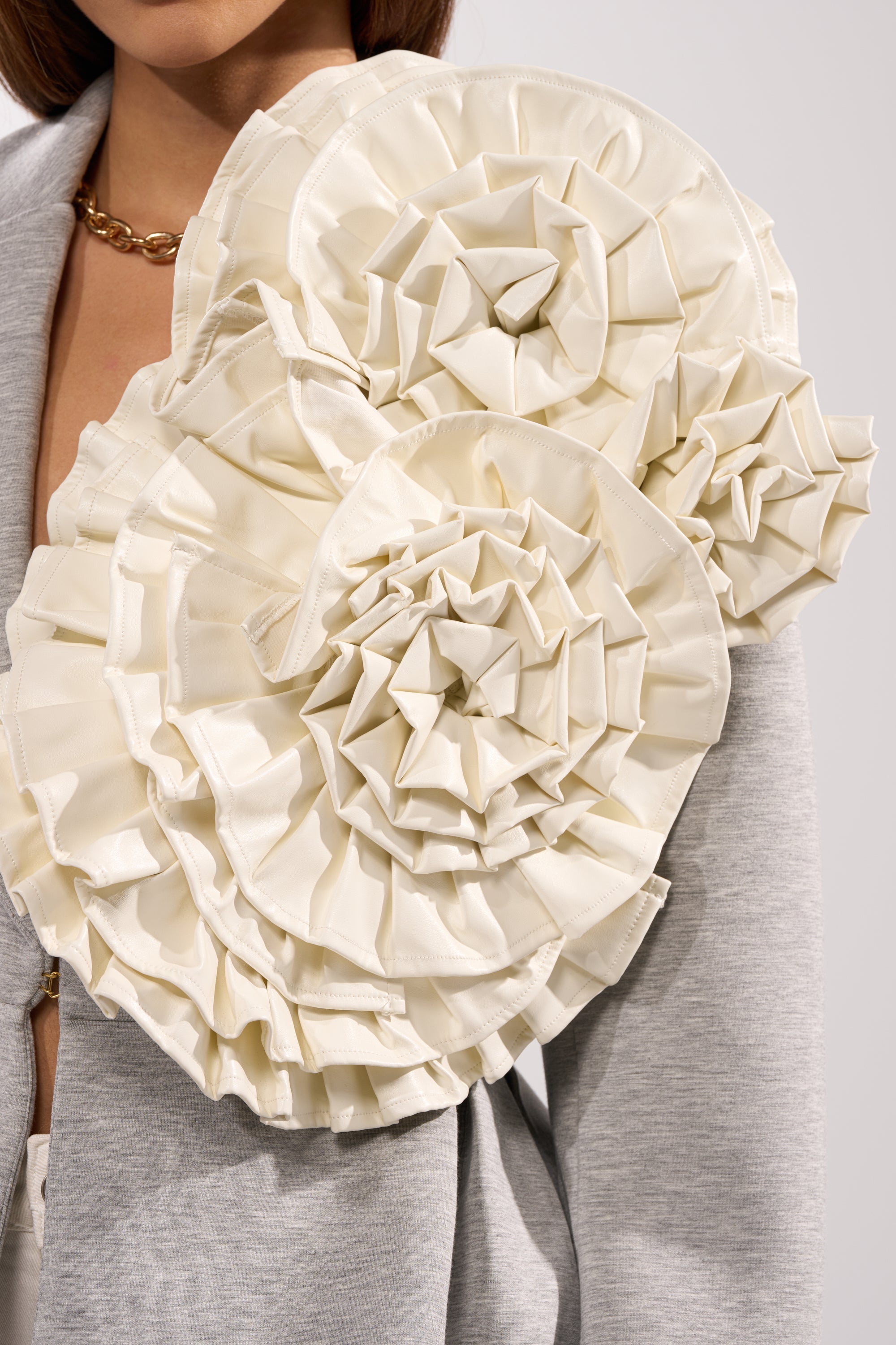 A person wears a grey jacket styled with the BLOOMING PU OVERSIZED BROOCH—large, ruffled cream fabric flowers on the shoulder—and a chunky gold chain necklace. 1