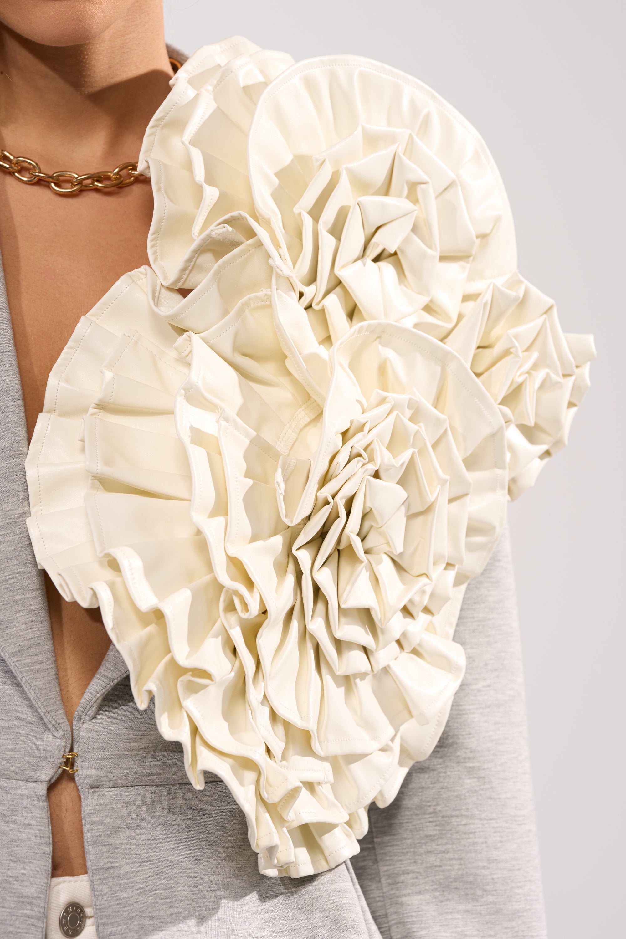 A person wears a grey blazer accented with a chunky gold chain necklace and the BLOOMING PU OVERSIZED BROOCH—an eye-catching, cream-colored floral accessory—visible from the neck down. 2